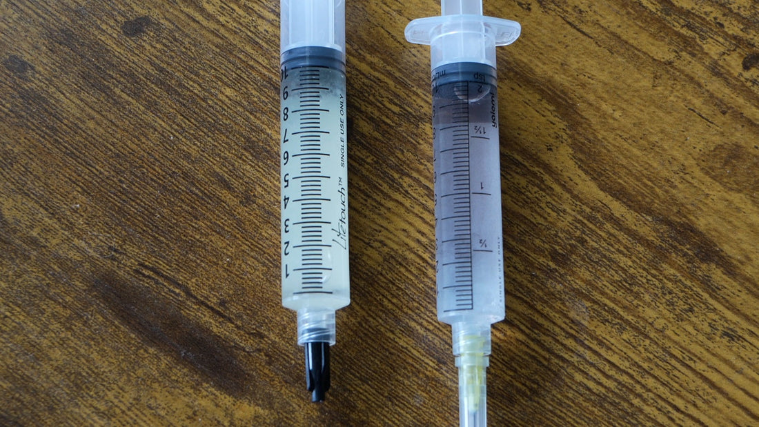 A liquid culture syringe and a spore syringe lying side by side on a wooden tabletop.